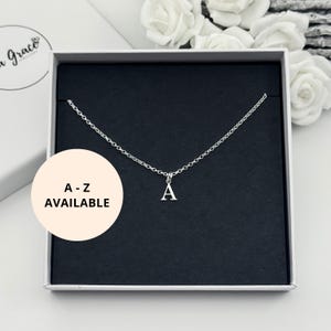 May include: A delicate silver chain necklace featuring a pendant in the shape of the letter "A". The necklace is displayed in a white gift box with a black interior. The text "A - Z AVAILABLE" is on a round sticker.