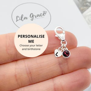 Peut inclure: Un charm en argent avec un fermoir mousqueton, un cœur avec la lettre « L » et une pierre précieuse ronde, rouge foncé. Le charm est tenu dans une main. L'image comprend le texte « Personnalisez-moi » et « Choisissez votre lettre et votre pierre de naissance ».