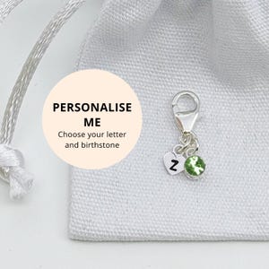 Puede incluir: Un charm de plata con un cierre de mosquetón, un corazón con la letra "Z" y una piedra preciosa verde. El charm se muestra sobre un fondo de tela blanca. También se ve el texto "PERSONALÍZAME" y "Elige tu letra y piedra de nacimiento".