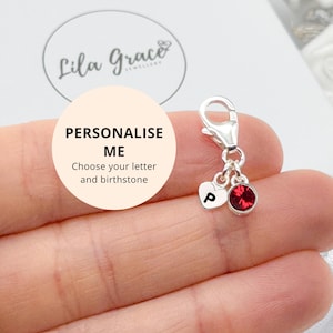 May include: A silver charm featuring a lobster clasp, a small heart-shaped pendant engraved with the letter P, and a round red crystal birthstone. The charm is displayed with text that reads Lila Grace Jewellery and PERSONALISE ME.
