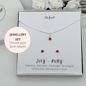Könnte beinhalten: Eine weiße Schachtel mit einem roten Rubin-Geburtstags-Halskette und Ohrring-Set. Die Schachtel trägt den Text "Lila Grace" und "JEWELLERY SET Choose your Birth Month". Die Schachtel zeigt auch den Text "JULY - RUBY Success. Passion. Courage. Strength Sterling Silver with Swarovski Crystals".
