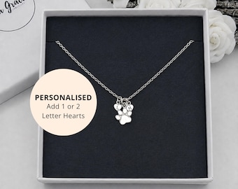 May include: A silver-toned necklace featuring a paw print charm and two small heart-shaped charms engraved with the letters L and M. The necklace is presented in a square white box with a black insert. Text on the box reads: PERSONALISED Add 1 or 2 Letter Hearts.