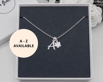 May include: A silver-toned necklace featuring a delicate chain with two charms: a capital letter A and a small paw print. The jewelry is presented in a square box with a dark gray insert. A circular overlay displays the text A - Z AVAILABLE.