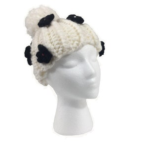 May include: White knitted beanie with a black pom pom and black flower embellishments.