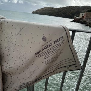 May include: A cream-coloured nautical map, labelled "The Scilly Isles", is draped over a metal railing. The map features detailed lines and text, with a dark blue print. The background shows a body of water and a cloudy sky.
