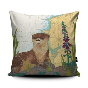 May include: A decorative cushion featuring a brown and tan otter with a floral pattern on its fur. The otter is sitting on rocks with green plants and a purple flower in the background. The background is a nautical chart with the text "Barnstaple" and "Bideford Bay".