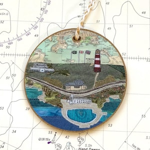 May include: A round wooden ornament featuring a stitched map of Plymouth, England. The map depicts a lighthouse, a harbour, and a coastline. The ornament is decorated with blue, green, and brown stitching.