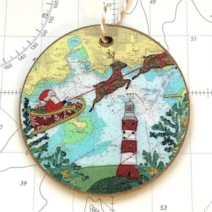 May include: Circular Christmas decoration depicting Santa's sleigh and reindeer flying over a nautical chart, with a red and white lighthouse. The ornament has a beige edge and a cream ribbon for hanging. The diameter is approximately 8 cm.