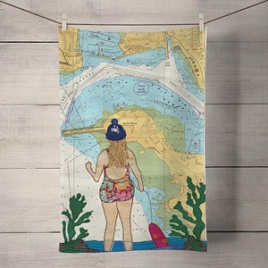 May include: A colourful tea towel with a nautical map design featuring a woman in a swimsuit and a blue beanie holding a surfboard. The map shows the area around Cattewater, England.