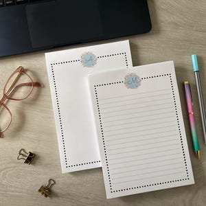 May include: Two white notepads with floral accents and initial letters, one lined and one blank, sit on a wooden surface. A laptop, glasses, binder clips, and pens are also present. The notepads have dotted borders.