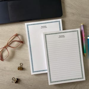 May include: Two white notepads with blue dotted borders and the word "Notes" at the top. A pair of rose-tinted glasses, two gold binder clips, a pen, a highlighter, and a laptop are on the wooden surface.