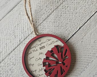 May include: A red and white wooden ornament with a cheerleading megaphone design. The ornament is personalized with the names of the cheerleaders, the coach's name, and the year 2025.