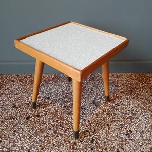 May include: A small, square side table with a light wood frame and legs. The tabletop features a white, textured surface. The legs are tapered and have dark metal tips. This vintage table is a functional and decorative piece.