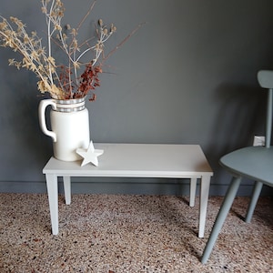 Peut inclure: Un banc rectangulaire en bois gris clair doté de quatre pieds droits. Sur le banc se trouve une cruche en céramique blanche avec un bord à carreaux gris et blanc, remplie de branches brunes séchées, et un petit ornement en forme d'étoile.