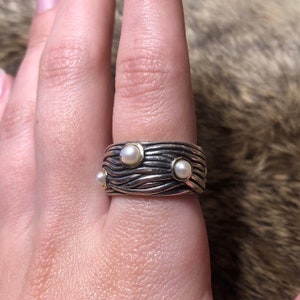 Könnte beinhalten: Ein Silber- und Goldring mit drei weißen Perlen, die in die Band gefasst sind. Die Band ist mit wellenförmigen Linien strukturiert.