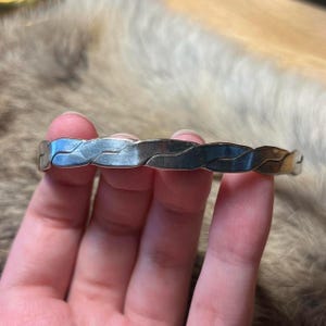 May include: A silver bangle bracelet with a braided pattern. The bracelet is made of a shiny metal and has a simple, elegant style. The bracelet is approximately 6.35 cm in diameter.