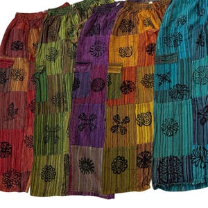Op de afbeelding: Een collectie van vijf losvallende broeken met een patchworkontwerp met verticale strepen en zwarte bloemen- en zonmotieven. Elke broek heeft een elastische tailleband met een trekkoord en een zijzak op het bovenbeen.