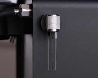 Puede incluir: Primer plano de una máquina de espresso negra con una herramienta de distribución de metal plateado. La herramienta tiene un mango texturizado y agujas finas. Se ven los componentes de la máquina.