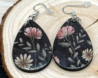 May include: A pair of teardrop-shaped wooden earrings with a floral print. The earrings are black with pink and white flowers and gold accents.