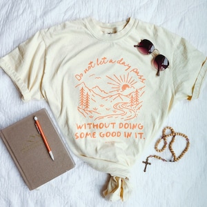 May include: A light yellow t-shirt with an orange graphic of a mountain scene and the text "Do not let a day pass without doing some good in it."