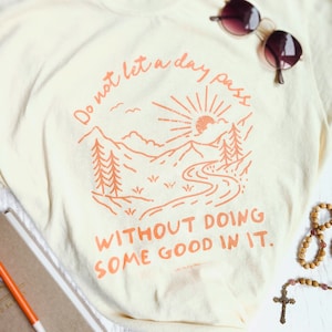 May include: A cream-coloured t-shirt with an orange graphic of a mountain range, sun, and river. The text "Do not let a day pass without doing some good in it." is printed on the shirt.