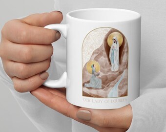 May include: White ceramic mug with a graphic of Our Lady of Lourdes appearing to a young girl in a cave. The graphic is surrounded by a gold and white floral design. The text "Our Lady of Lourdes" is printed on the bottom of the mug.