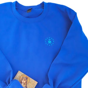 May include: A royal blue sweatshirt with a small embroidered design on the chest. The design features a blue and white 'M' inside a circle of stars. The sweatshirt is folded and laid flat on a white background.