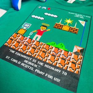 May include: Green t-shirt with a pixelated video game design featuring a character holding a cross. The text reads "Always be close to Jesus. World is Level 200." and "The Eucharist is the highway to heaven. St. Carlo Acutis, pray for us!"