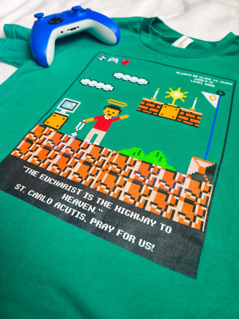 May include: Green t-shirt with a pixelated video game design featuring a character holding a cross. The text reads "Always be close to Jesus. World is Level 200." and "The Eucharist is the highway to heaven. St. Carlo Acutis, pray for us!"