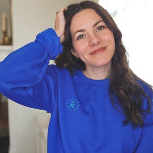 May include: A woman wearing a royal blue sweatshirt with a light blue embroidered design of a star and the letter "M" surrounded by stars.