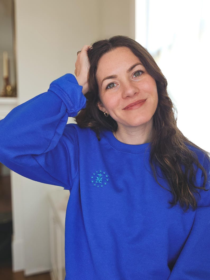 May include: A woman wearing a royal blue sweatshirt with a light blue embroidered design of a star and the letter "M" surrounded by stars.