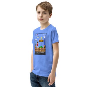 May include: A light blue t-shirt for kids featuring a pixelated image of a video game character standing on a platform. The text on the shirt reads "THE STRUGGLE IS THE NECESSARY TO REACH HEAVEN. 12 CHALLENGES, PRAY FOR HIM."