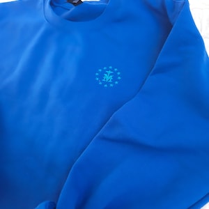 May include: A royal blue sweatshirt with a small embroidered design of a circle with stars and a cross inside. The design is in a light blue colour.