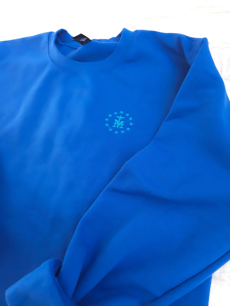 May include: A royal blue sweatshirt with a small embroidered design of a circle with stars and a cross inside. The design is in a light blue colour.