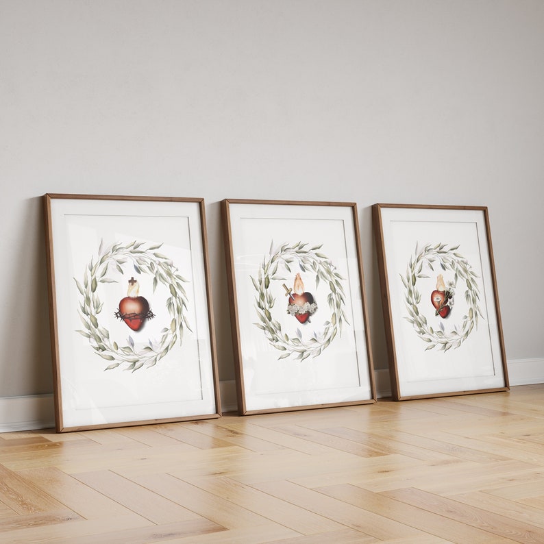 May include: Three framed art prints. Each print features a heart with a crown of thorns, a flame, and a cross, encircled by a green leaf wreath. The frames are brown, and the prints lean against a white wall on a wooden floor.