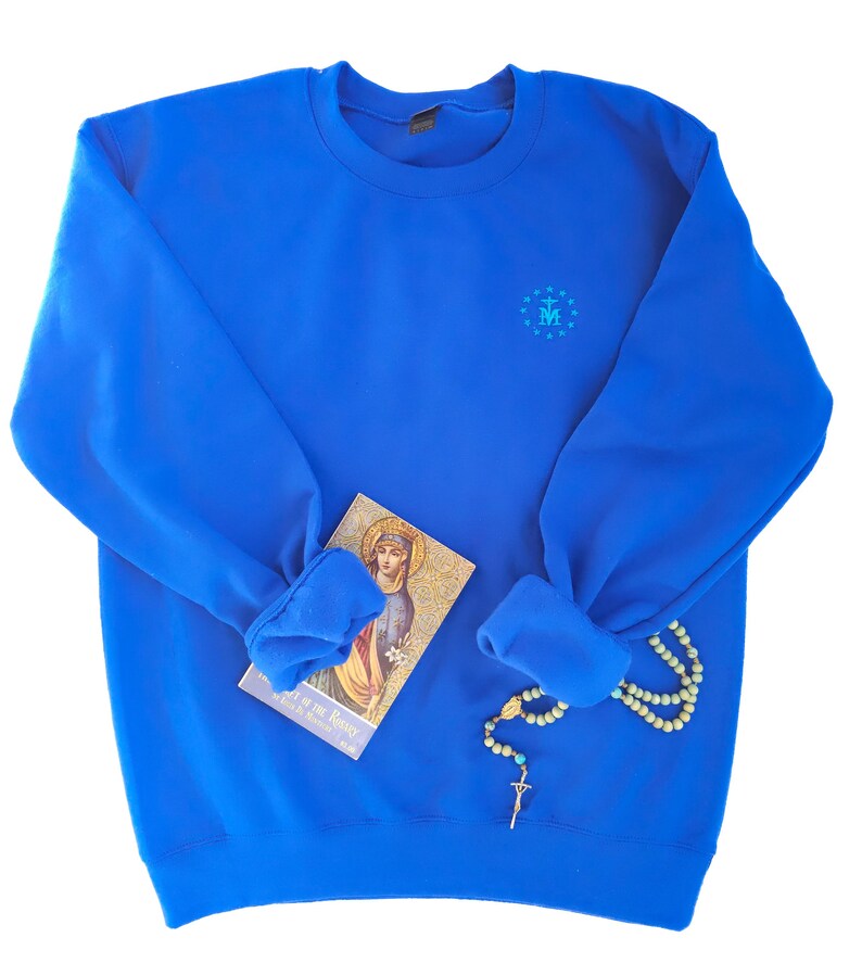 May include: A royal blue sweatshirt with a small embroidered design on the chest. The design features a blue and white 'M' inside a circle of stars. The sweatshirt is folded and laid flat on a white background.