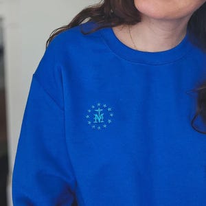 May include: A royal blue sweatshirt featuring a light blue embroidered emblem of the Virgin Mary. The emblem is surrounded by a circle of small stars.