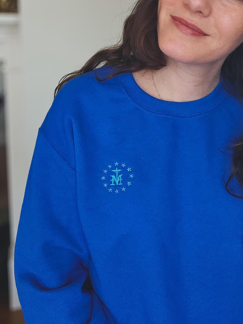 May include: A royal blue sweatshirt featuring a light blue embroidered emblem of the Virgin Mary. The emblem is surrounded by a circle of small stars.
