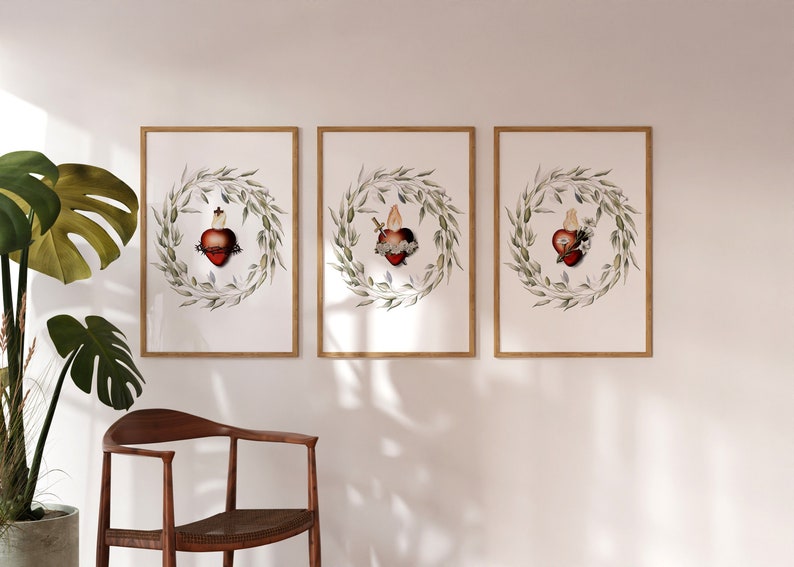 May include: Three framed art prints. Each print features a red heart with a crown of thorns, encircled by a green leaf wreath. The prints are displayed on a white wall. A wooden chair and a potted plant are in the foreground.
