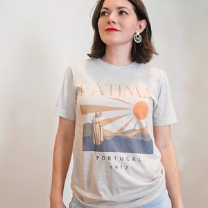 Op de afbeelding: Een lichtgrijs T-shirt met een grafische print van een vrouw in een witte jurk die voor een zon en bergen staat. De tekst "FATIMA PORTUGAL 1917" is onder de afbeelding gedrukt.