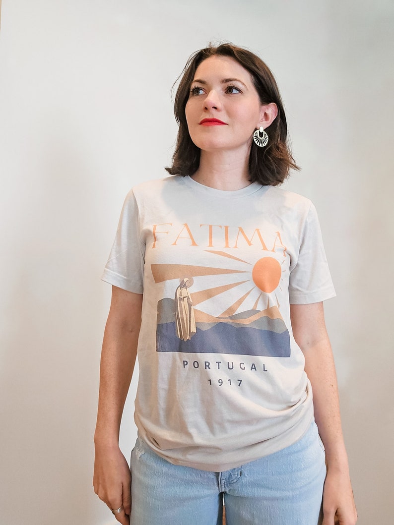 Op de afbeelding: Een lichtgrijs T-shirt met een grafische print van een vrouw in een witte jurk die voor een zon en bergen staat. De tekst "FATIMA PORTUGAL 1917" is onder de afbeelding gedrukt.