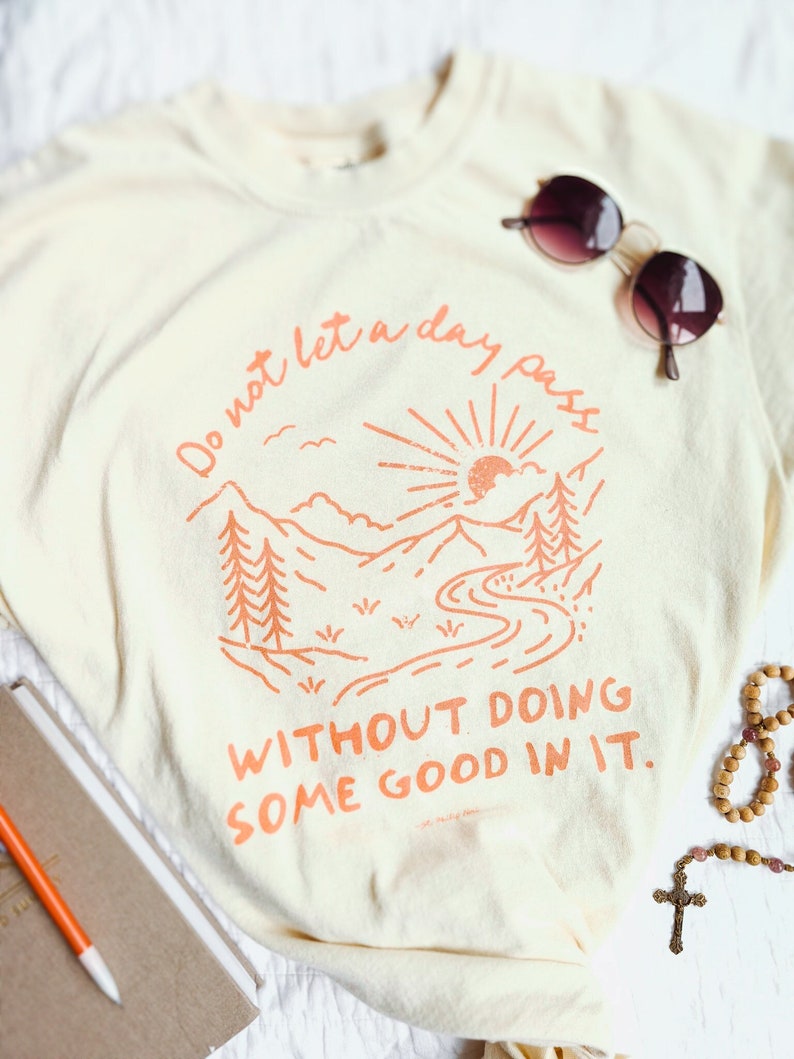 May include: A cream-coloured t-shirt with an orange graphic of a mountain range, sun, and river. The text "Do not let a day pass without doing some good in it." is printed on the shirt.