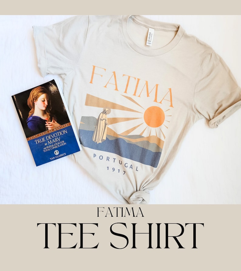 Op de afbeelding: Een beige T-shirt met het woord "FATIMA" in oranje op de voorkant. Het shirt heeft ook een afbeelding van een persoon in een gewaad, een zon en de woorden "PORTUGAL 1917". Een boek met de titel "TRUE DEVOTION TO MARY" ligt op het shirt.