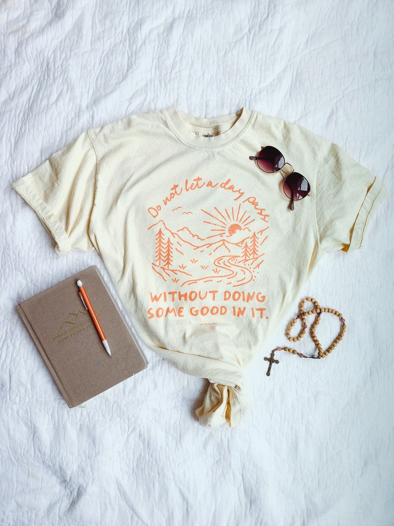 May include: A light yellow t-shirt with an orange graphic of a mountain scene and the text "Do not let a day pass without doing some good in it."