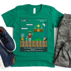 May include: Green t-shirt with a pixelated video game design featuring a character holding a cross. The text reads "The Eucharist is the highway to heaven. St. Carlo Acutis, pray for us!"