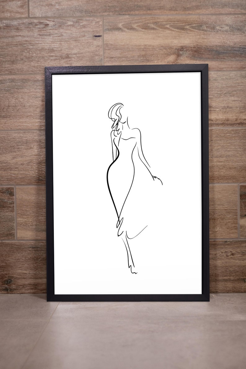May include: A framed black and white line art print. The artwork depicts a woman in a flowing dress, rendered with simple, elegant lines. The minimalist design is set against a white background, framed in a black border.