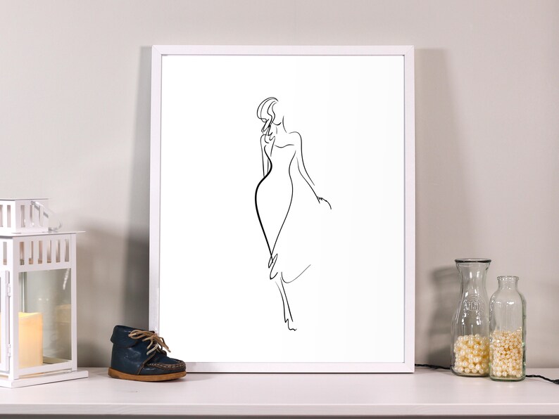 May include: A framed minimalist line art print of a woman in a dress. The artwork is a simple black outline on a white background, set within a white frame. The print is displayed on a white surface with decorative items.