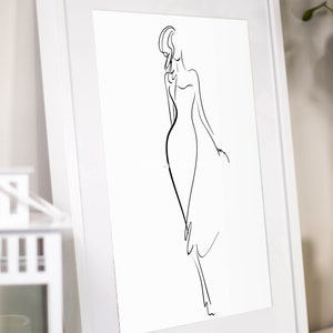 May include: A framed art print featuring a minimalist line drawing of a woman in a flowing dress. The artwork is rendered in black against a white background, encased in a white frame. The image is leaning against a wall.
