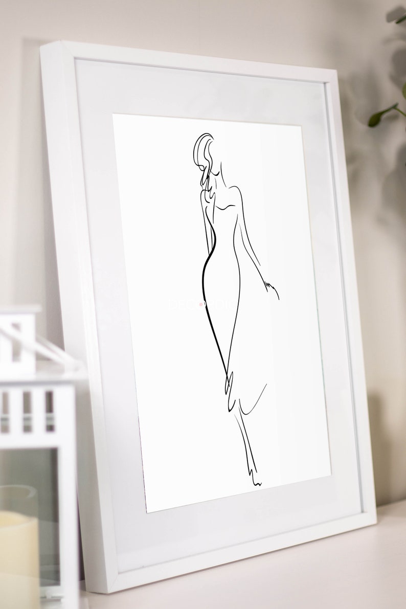 May include: A framed art print featuring a minimalist line drawing of a woman in a flowing dress. The artwork is rendered in black against a white background, encased in a white frame. The image is leaning against a wall.