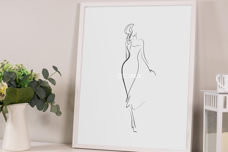 May include: A framed minimalist art print featuring a black line drawing of a woman in a flowing dress. The artwork is set against a white background within a white frame. The text "DECORIDIC" is visible on the artwork.